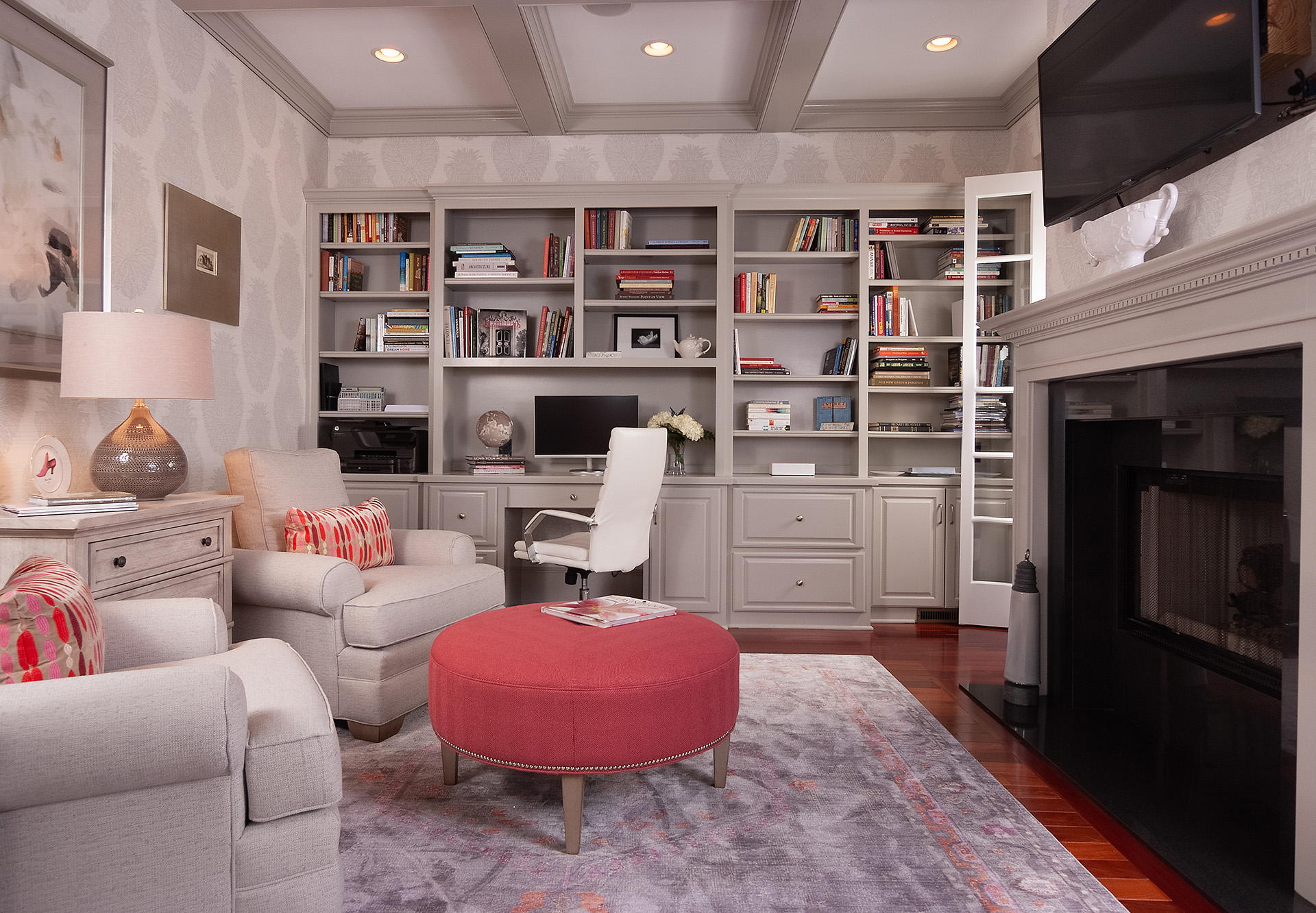 Built-in shelving with a desk in a basement with a coffered ceiling. Grey colour scheme with pink ottoman and throw pillows on armchairs.