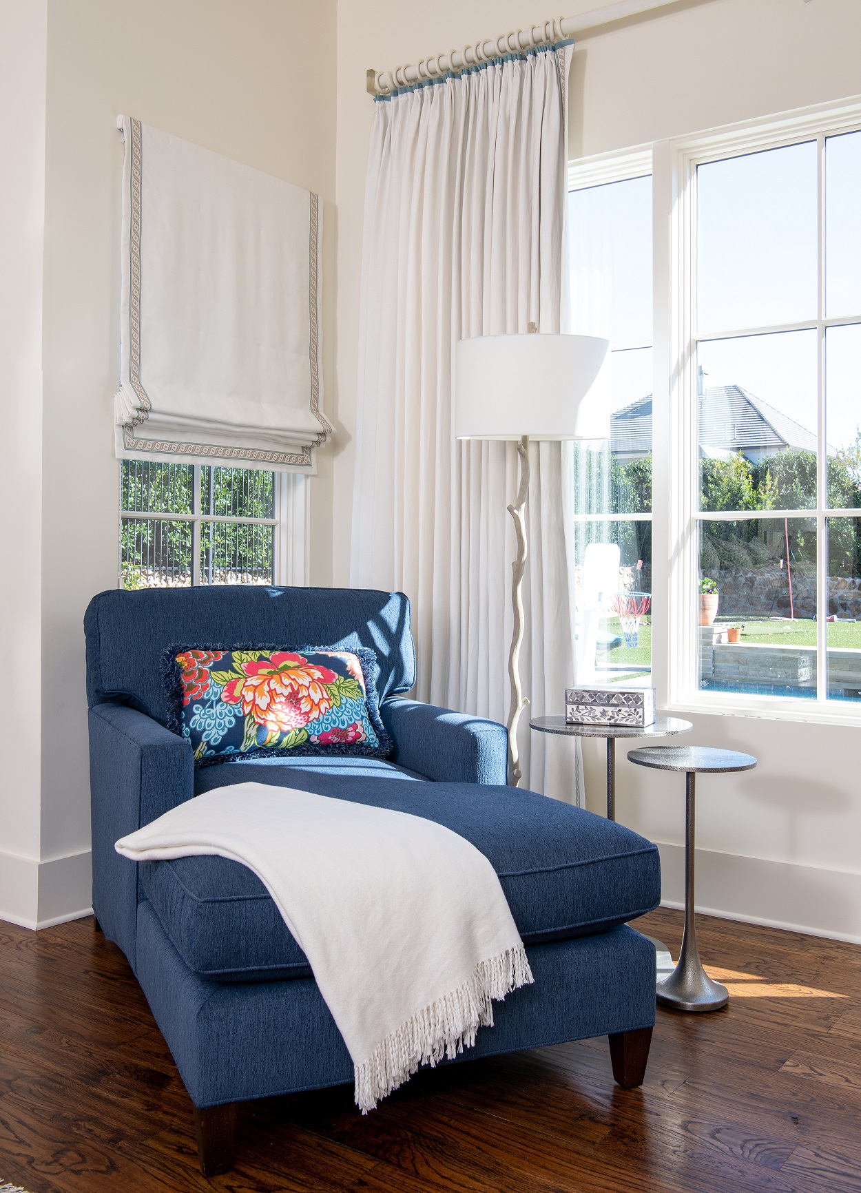 A blue upholstered chaise in a transitional style is the focal point of the room. The chaise is positioned in front of a window, with a white roman shade and drapes in another window. The floor is a deep brown hardwood, and the walls are white. A colourful floral kidney pillow and a white throw are placed on the chaise. Two drinks tables finished in brushed nickel are situated near the chaise, with a silver and white floor lamp nearby. The room is well-lit and the blue chaise adds a pop of color to the space.