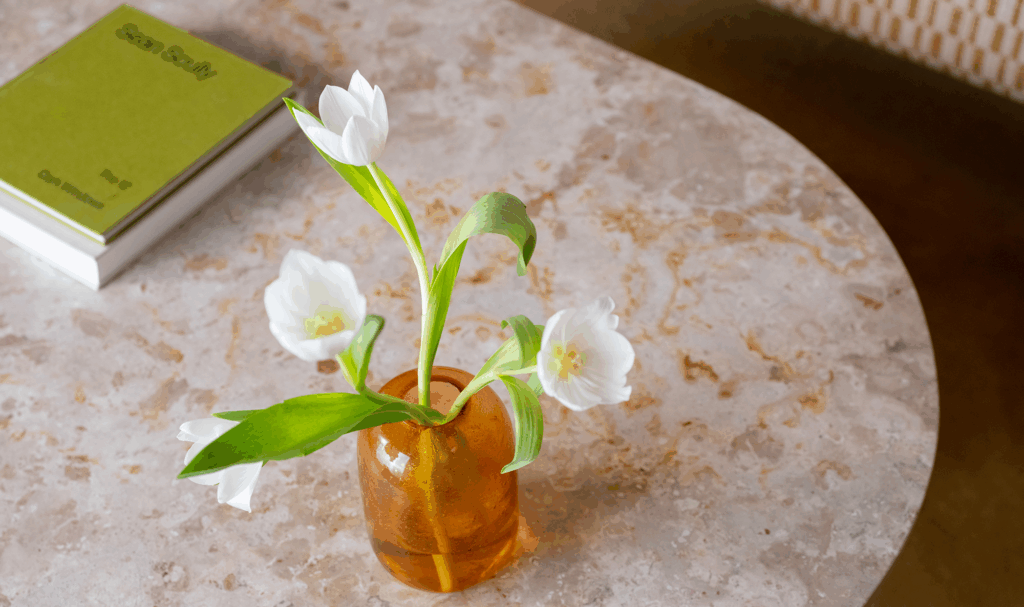 Close-up of marble coffee table with amber glass vase and white tulips – natural stone design trend 2026.
